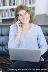 Smiling woman sitting on sofa at home using a laptop and speaking on phone 0LdKee