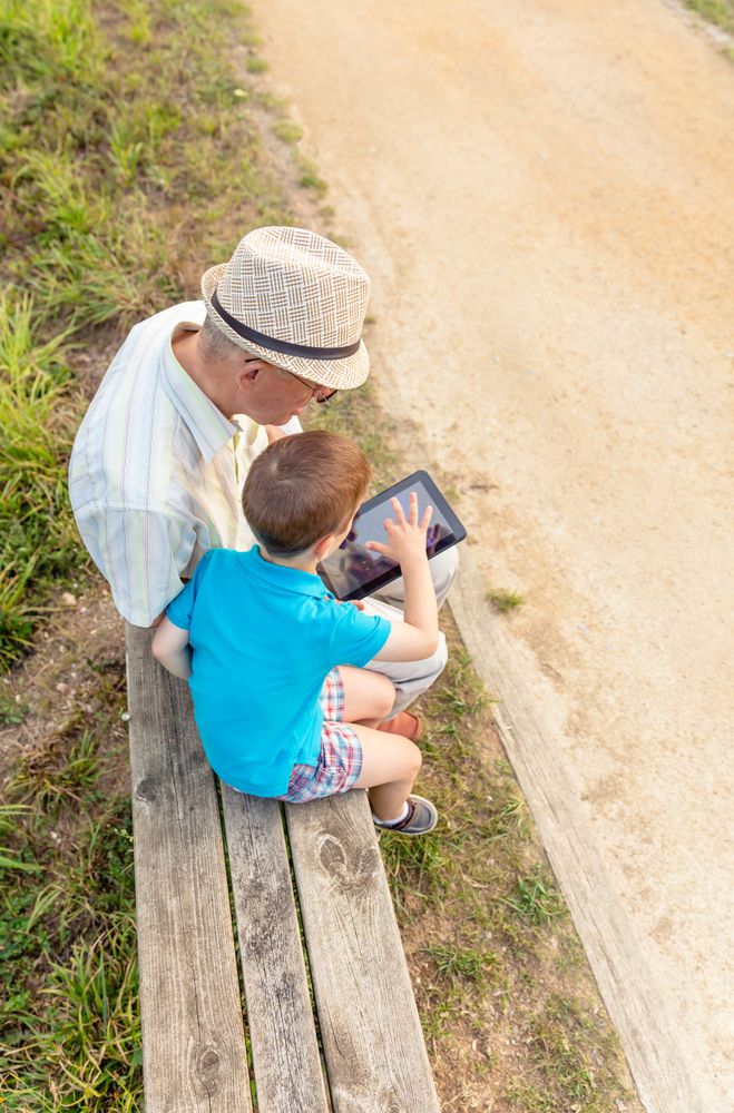 Grandchild and grandfather using a tablet outdoors on park bench, vertical