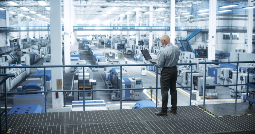 Experienced Male Engineer Standing on a Platform, Using Laptop Computer and Overlooking Production at a Modern Automated Electronics Manufacture with AI Support. Factory Executive at Work