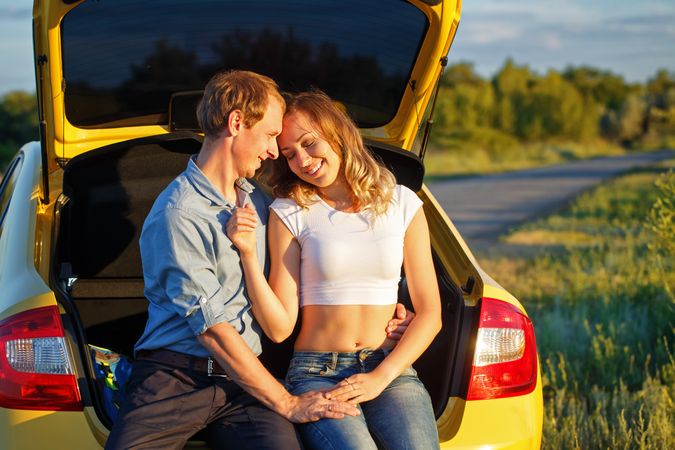 Couple sitting in back of broken down yellow car on side of the road