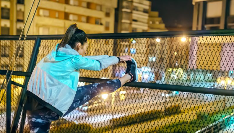 Brunette female runner stretching her legs over bridge railing before training in city at night