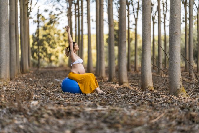 Adult pregnant lady sitting on big pilates ball in park during sunny day