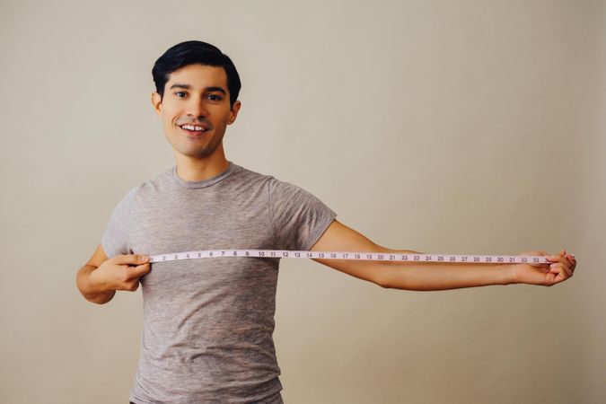 Smiling Hispanic male holding out measuring tape out to his side in beige studio shoot