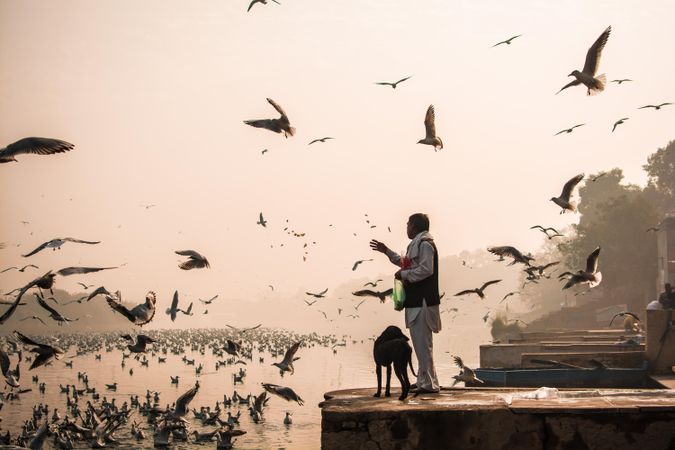 Side view of an Indian man in kurta standing by riverside with a dog while birds flying
