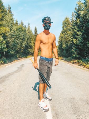 Topless man with facemask holding a baseball bat standing between trees