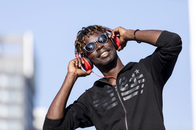 Smiling Black man wearing headphones & sunglasses standing on the street listening to music