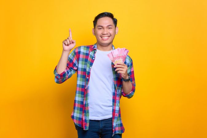 Asian man holding money he won and pointing upwards in studio shoot