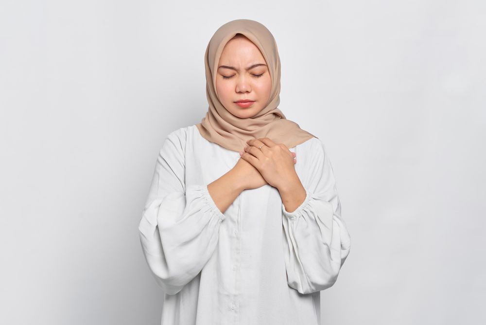 Sad Muslim woman in headscarf and light blouse looking down with hand over her heart