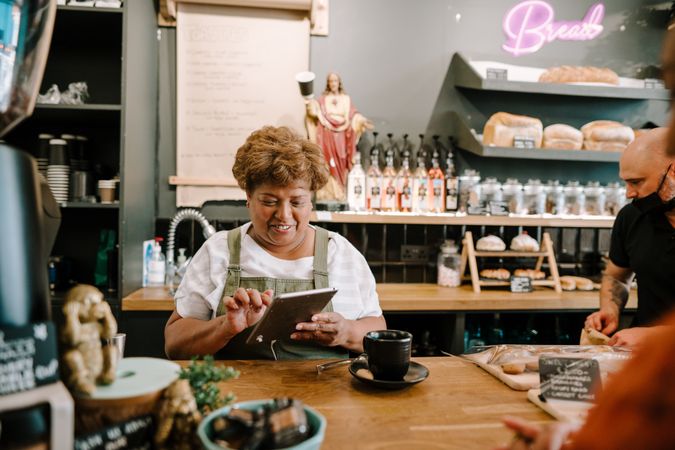 Smiling older Black woman ringing up order on digital tablet in coffee shop