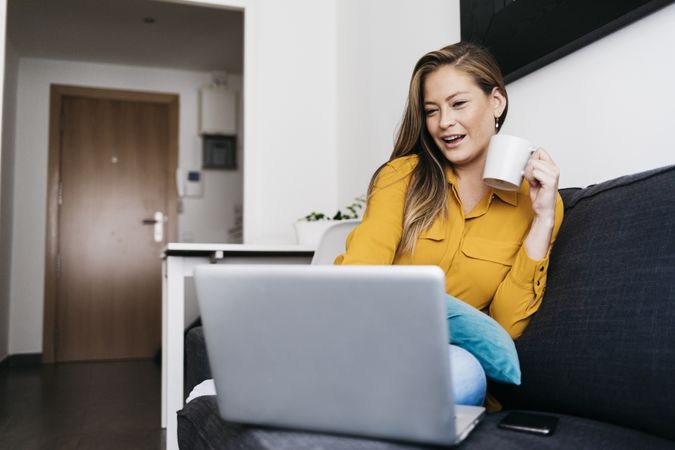 Happy long haired female sitting on sofa with laptop computer working remotely from home
