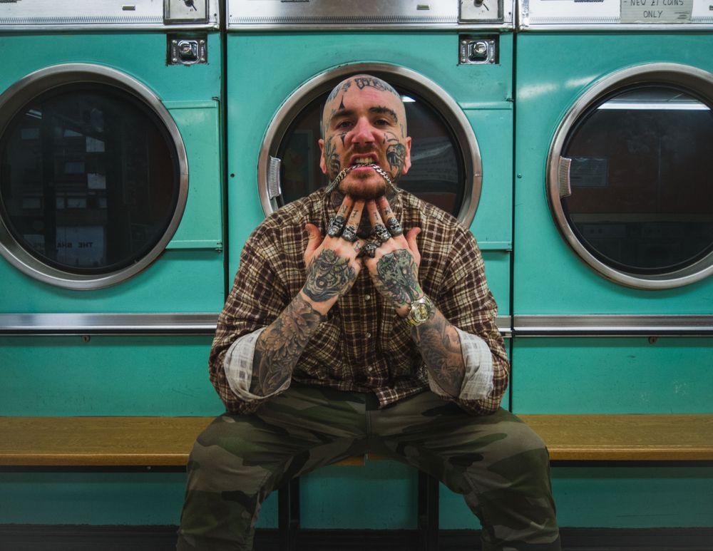 Edgy bald man with face tattoos in plaid shirt biting chain necklace in laundromat