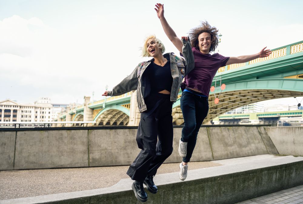 Male and female jumping and smiling on riverwalk with bridge in background