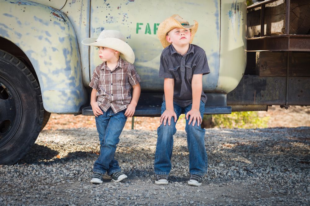 Two Boys Wearing Cowboy Hats Leaning Against an Antique Truck in a Rustic Setting