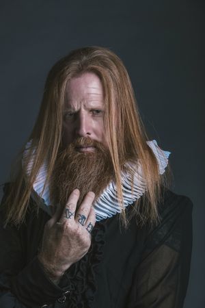 Confident adult red haired medieval magnate touching beard and looking at camera