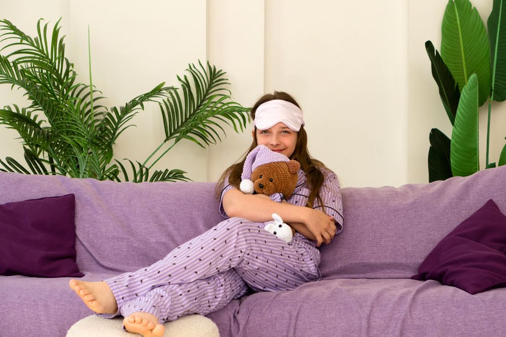 Girl cuddling teddy bear on couch with sleep mask.