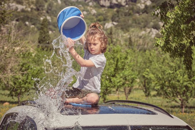 Cheerful boy pouring bucket of water on car