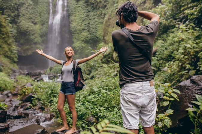 Excited young woman standing in front of a waterfall being photographed by her boyfriend