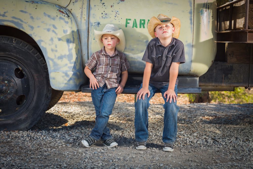 Two Boys Wearing Cowboy Hats Leaning Against an Antique Truck in a Country Setting