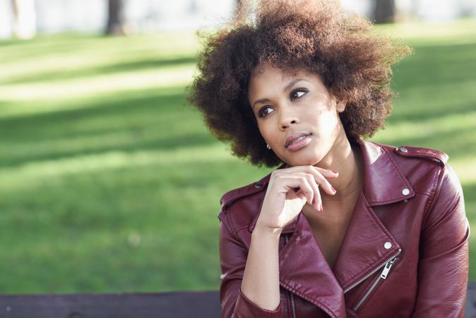 Contemplative woman sitting outside in red leather jacket on bright day with grass in background