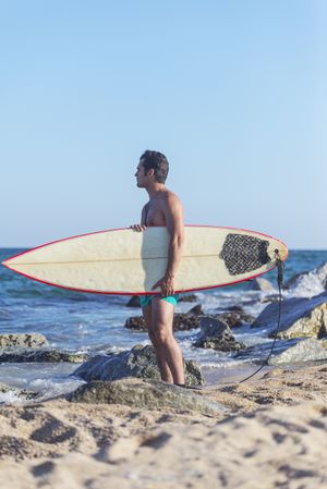 Male surfer standing on beach with board attached to his leg with leash on sunny day