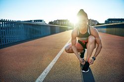 Happy athletic woman leaning down on bridge tying her shoes with sunflare