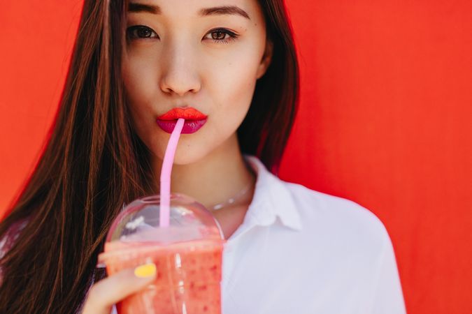 Close up of a young woman enjoying a fruit smoothie against a red wall