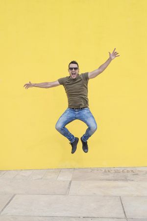 Joyful male standing outside jumping in front of yellow wall with open arms