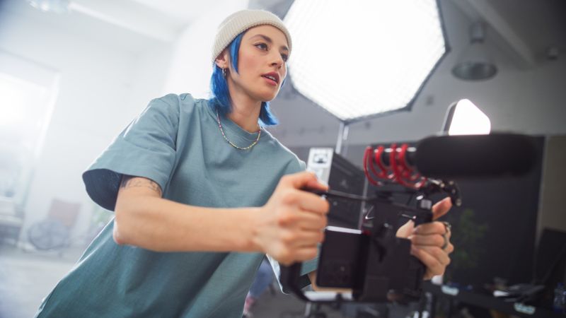 Focused Young Female Videographer With Blue Hair Adjusts Her Camera During An Indoor Film Shoot, Capturing The Essence Of Modern Filmmaking And Creative Production In A Professional Setting.