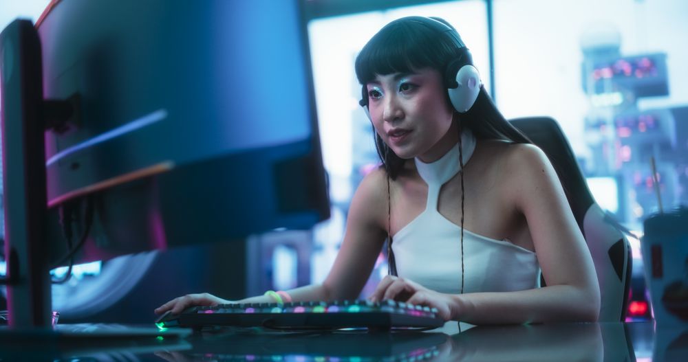 Enthusiastic Asian Woman in Headphones Talking with Friends Online on a Computer.