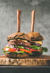 Two cheeseburgers skewered with knives, stacked with fresh vegetables, on wooden board