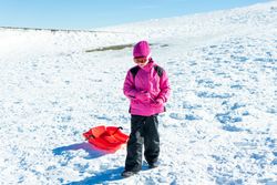 Child in pink snow suit tugging red sled behind her up a snowy hill