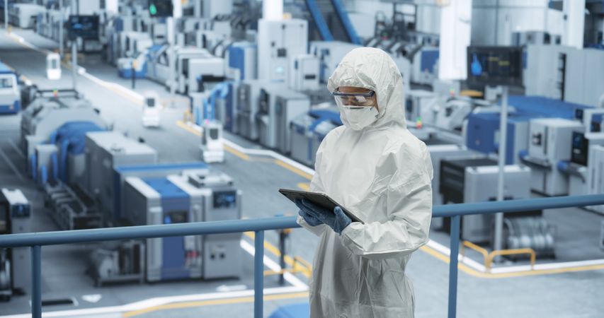 Female Technician in Protective Coveralls, Mask and Eyewear Using Tablet Computer at a Manufacturing Complex. Specialist Monitoring Conditions at a Modern Electronics Factory with Automated Robots