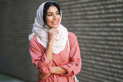Happy female in headscarf and pink trench coat standing confidently with with one hand to her chin