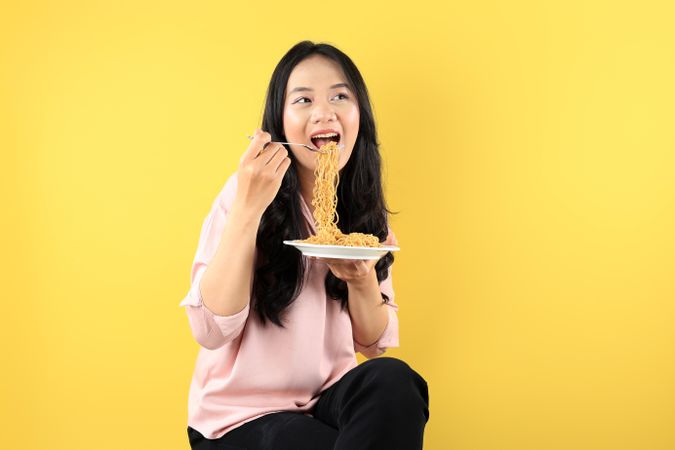 Young Asian Woman Holding A Plate of Fried Instant Noodle, Sitting on Yellow Background