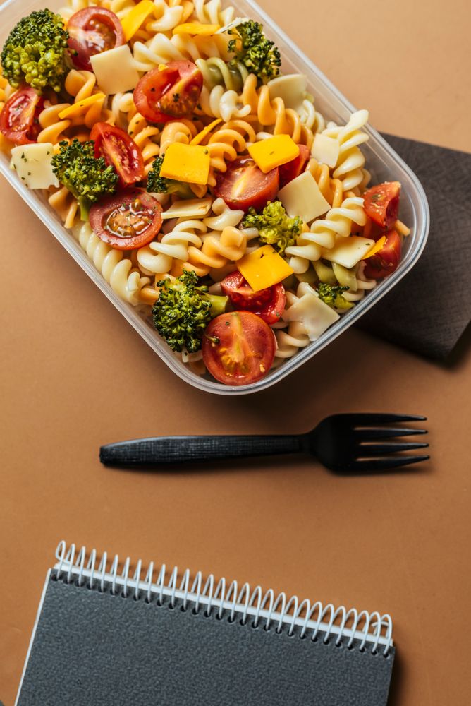 Lunchbox with pasta and plastic fork by notepad and pencils