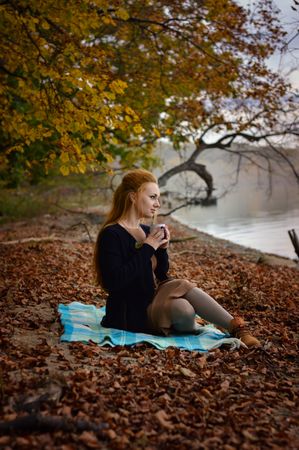 Woman holding a mug and sitting on autumn leaves beside waterside