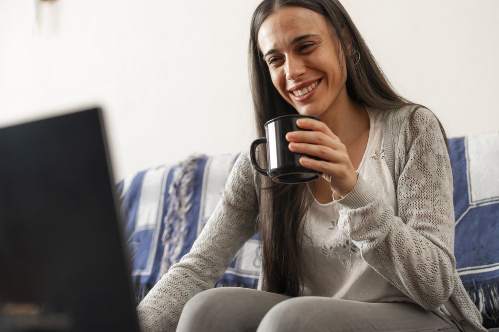 A woman enjoys a cup of tea, sitting on the sofa at home, while surfing the Internet from her laptop pc
