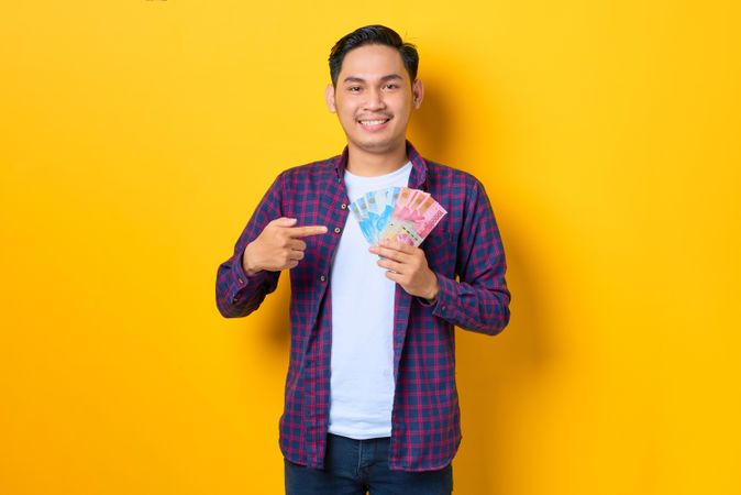 Happy young Asian man in plaid shirt pointing at banknotes