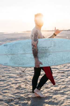 Tattooed man with surfboard under his arm making hang loose sign