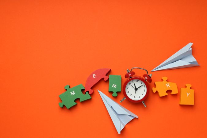 Puzzle pieces spelling “memory” on orange background with alarm clock and paper planes, copy space