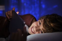 Cheerful boy with tablet lying on bed at night