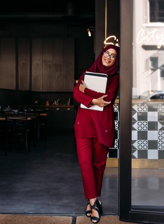 Woman wearing hijab holding laptop computer and looking at camera while standing at coffee shop