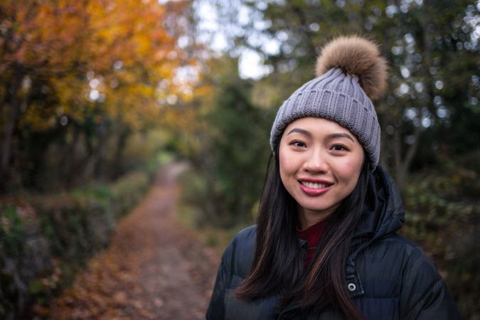 Smiling Asian woman in warm clothes standing in park with trees and stony fence