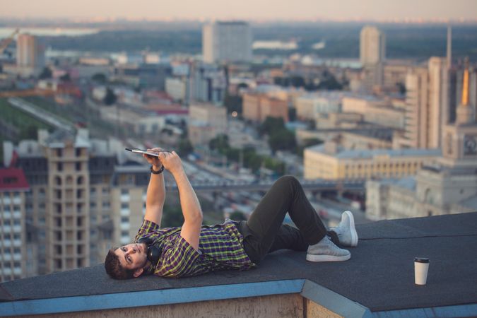 Male lying back looking at tablet, with headphones and takeaway coffee cup on rooftop