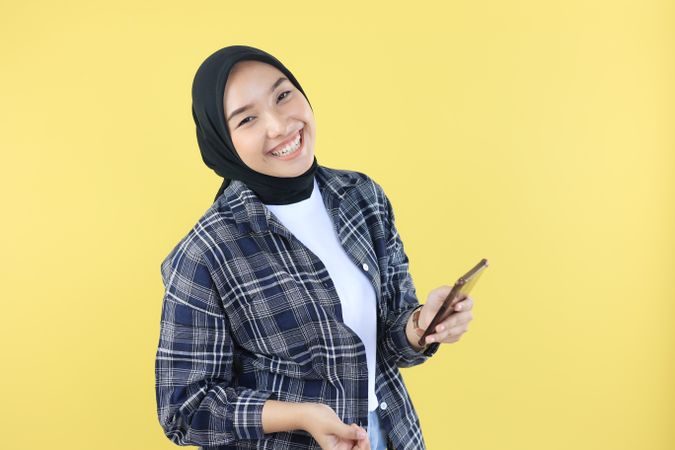Asian Young Woman Wearing Hijab Cheerful using a Smartphone Isolated on Yellow Background