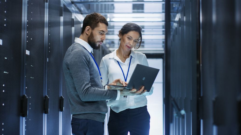 Male IT Specialist Holds Laptop and Discusses Work with Female Server Technician.