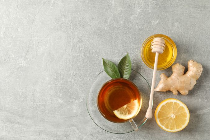 Cup of tea, leaves, ginger, lemon, honey and dipper on grey background, top view