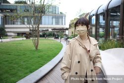 East Asian woman in beige coat with facemask standing near building bekAp5