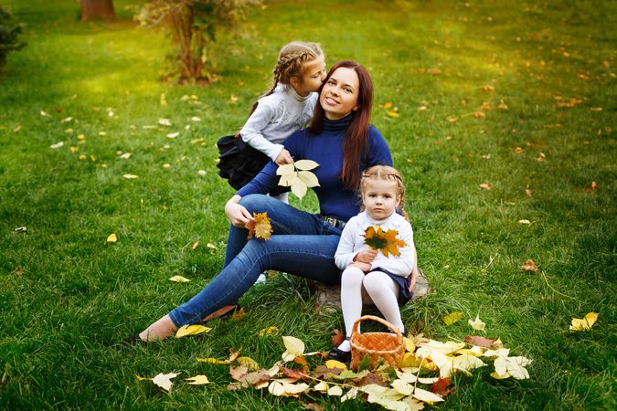 Two young blonde girls sitting with their mother in the park collecting leaves with basket