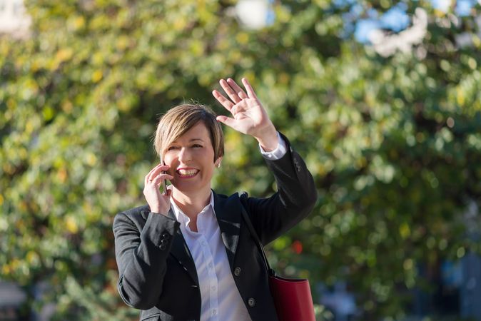 Businesswoman speaking on phone in front of hedge while waving